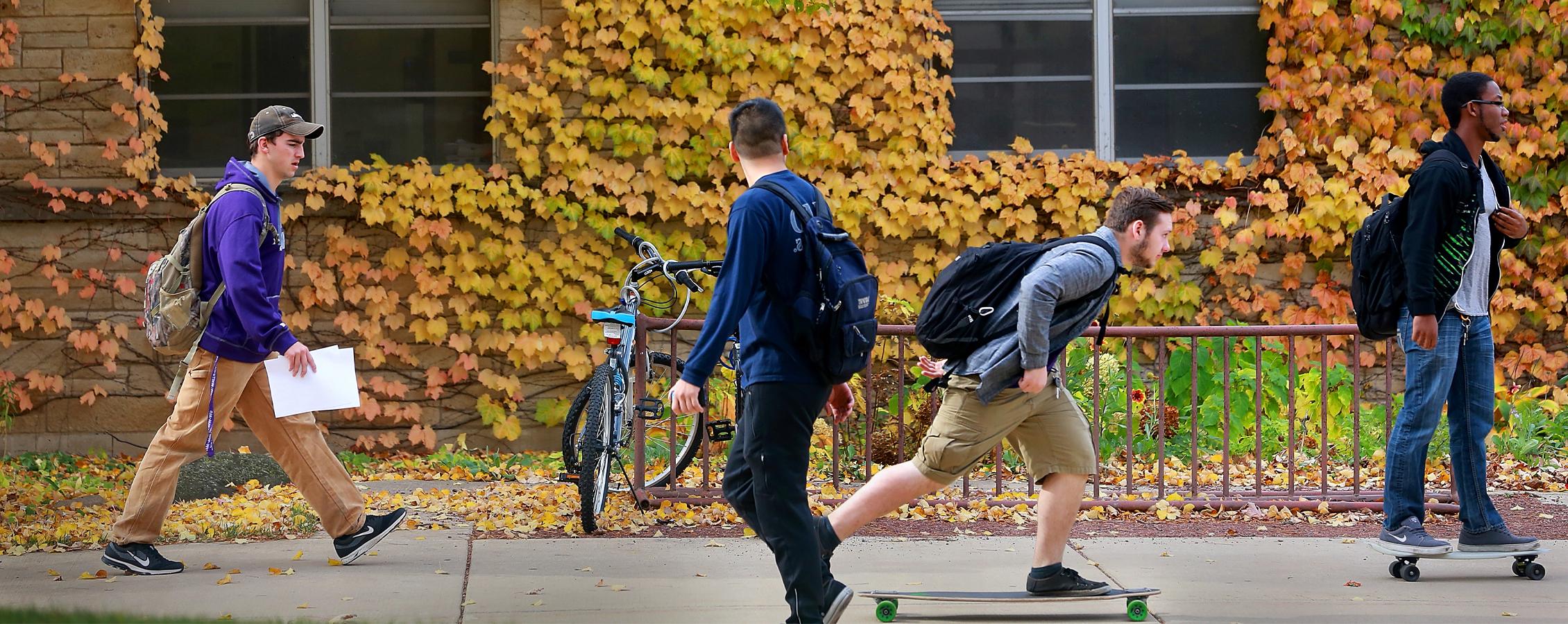 Students skateboard down a sidewalk with yellow foliage and fallen leaves in the background.